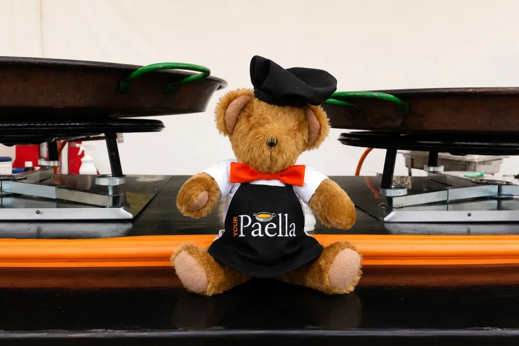 Your Paella teddy bear mascot sitting between two large paella pans on the Your Paella catering cart.